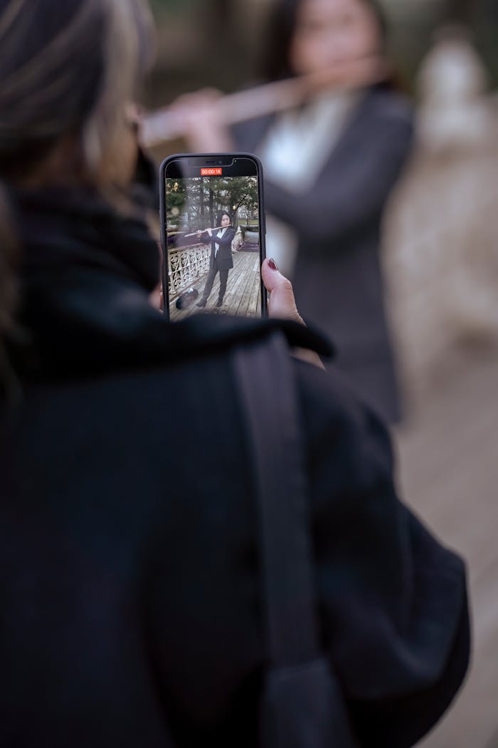 Home Woman recording a musician playing the flute outdoors, captured on a smartphone.